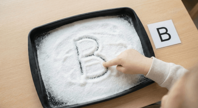 Child tracing letter B in salt tray for pre-writing practice