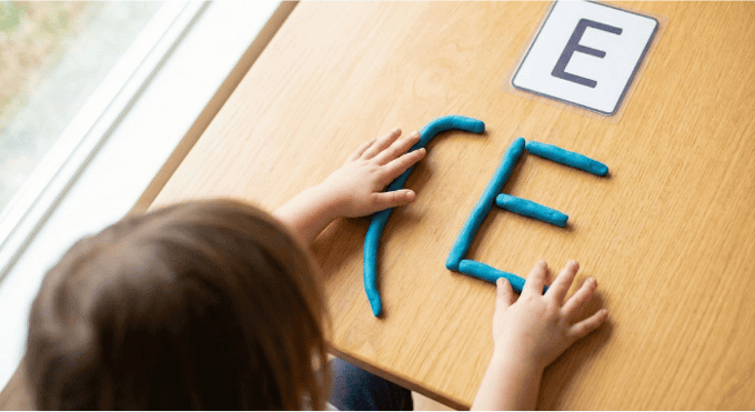 Preschooler forming letter E shape with blue playdough on table