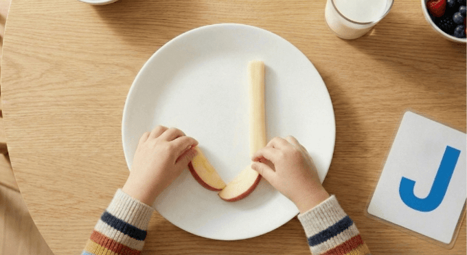 Preschooler forming letter J shape with cheese stick and apple slices for edible learning activity