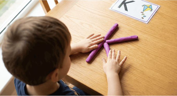Preschooler forming letter K shape with purple playdough on table