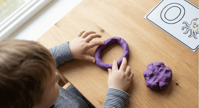 Preschooler forming letter O shape with purple playdough on table