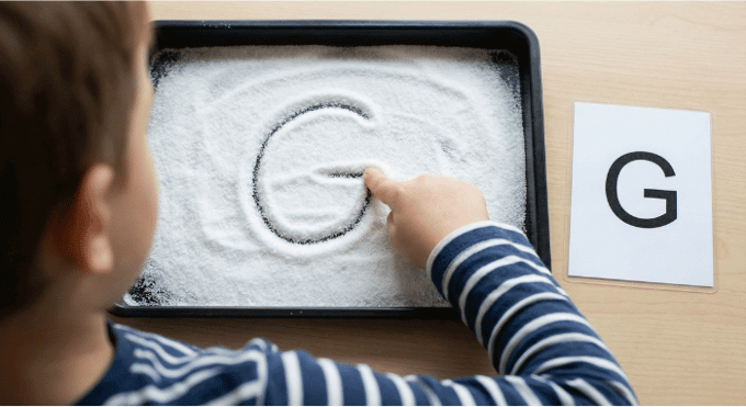 Child tracing letter G in salt tray for pre-writing practice
