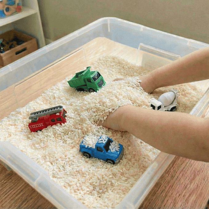 Preschool sensory bin activity with hands finding toy trucks in rice