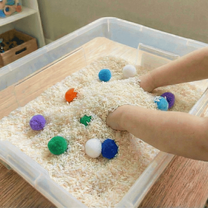 Preschool sensory bin activity with hands finding craft pom-poms in rice