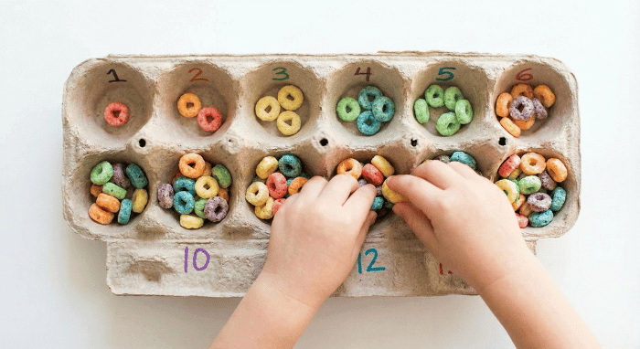 Egg carton with numbers written in each cup and colorful cereal pieces sorted by quantity for preschool counting activity